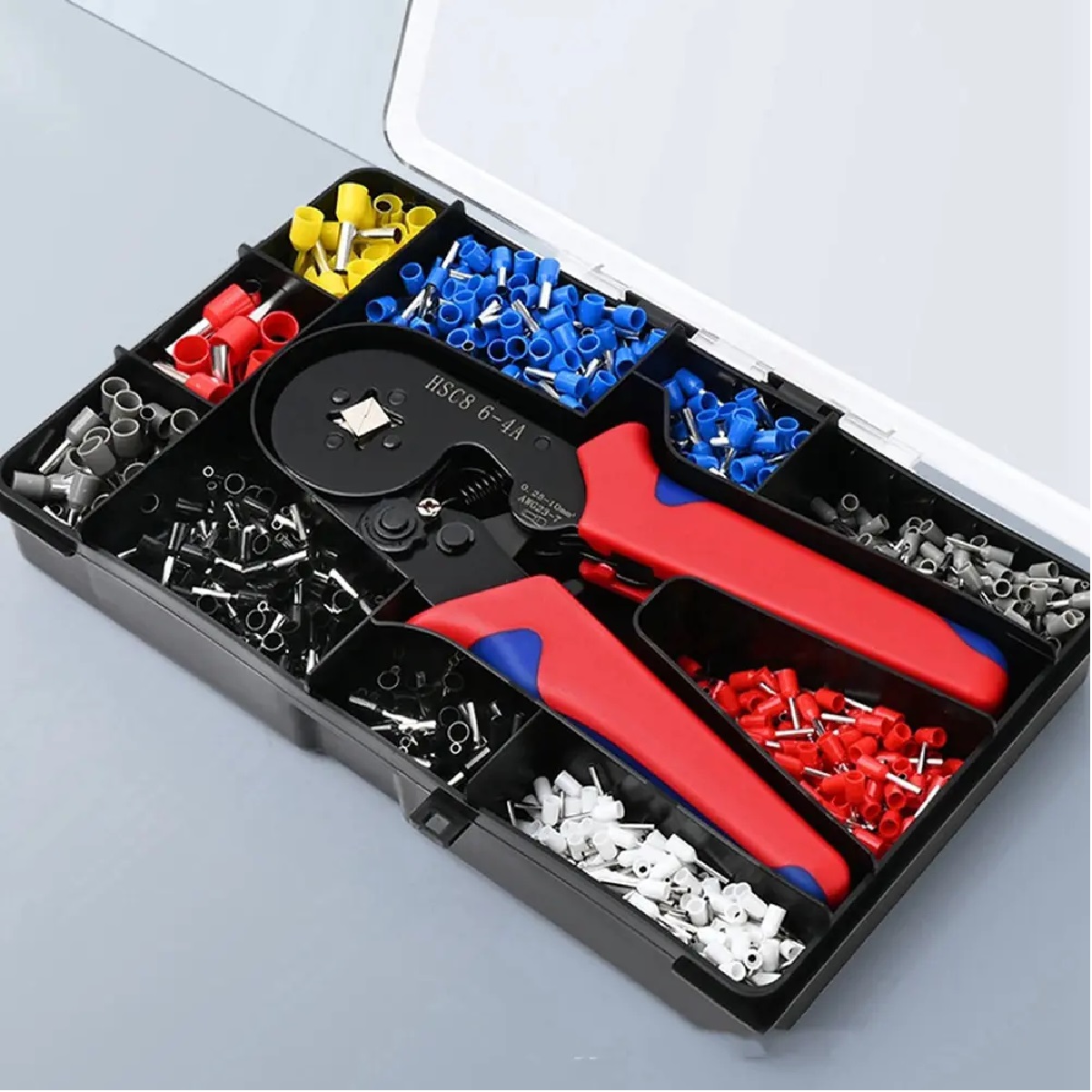 Pinza prensadora de terminales tubulares con trinquete, en caja organizadora negra con compartimentos para conectores amarillos, rojos, azules y blancos, sobre fondo gris claro.