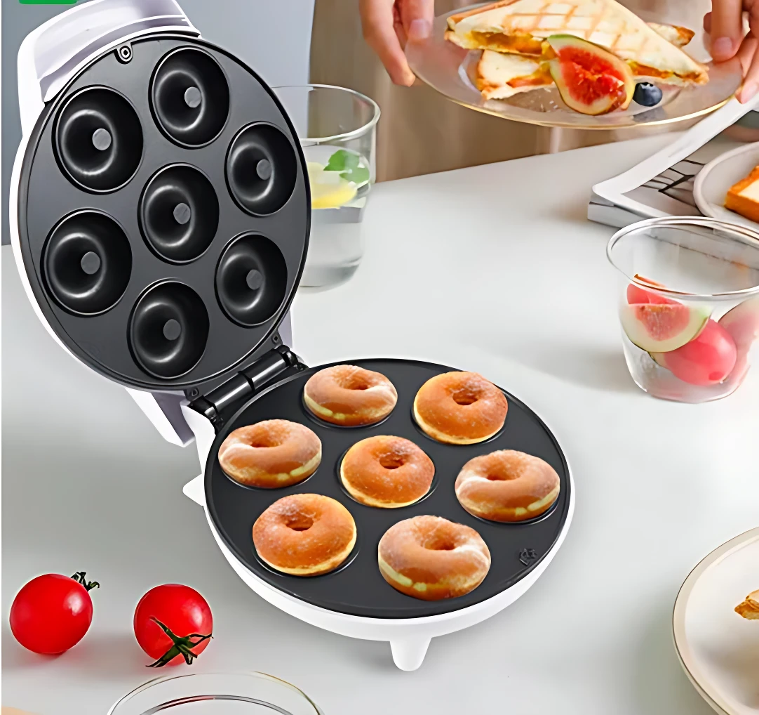 Máquina para hacer donas con siete moldes negros, abierta sobre mesa blanca, mostrando donas horneadas y accesorios de cocina al fondo.